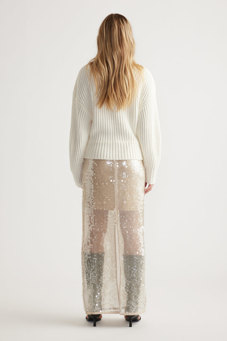 Woman wearing an off-white ribbed knit jumper paired with a shimmering sequin maxi skirt and black heels, shown from behind.