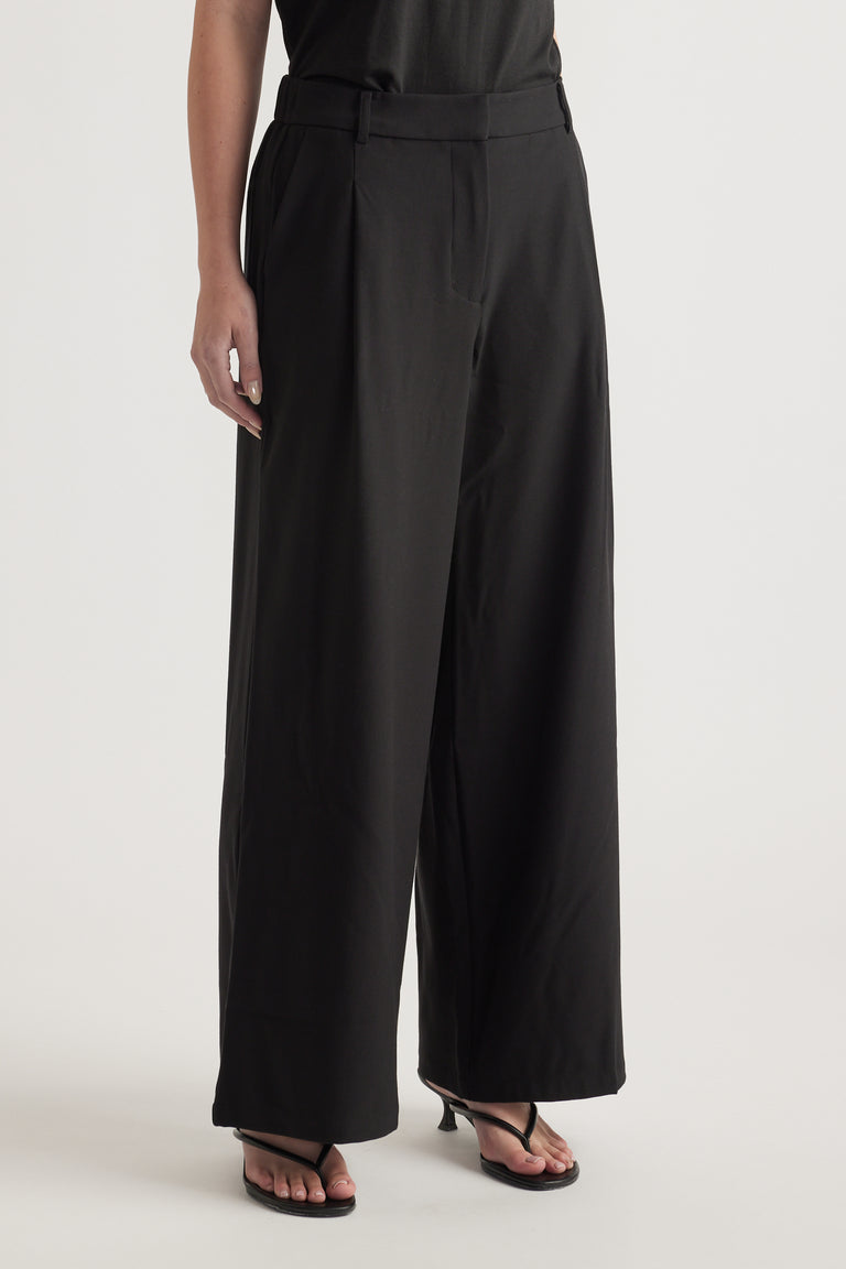 Woman wearing high rise black tailored wide leg pants with a zip fly and full length hem