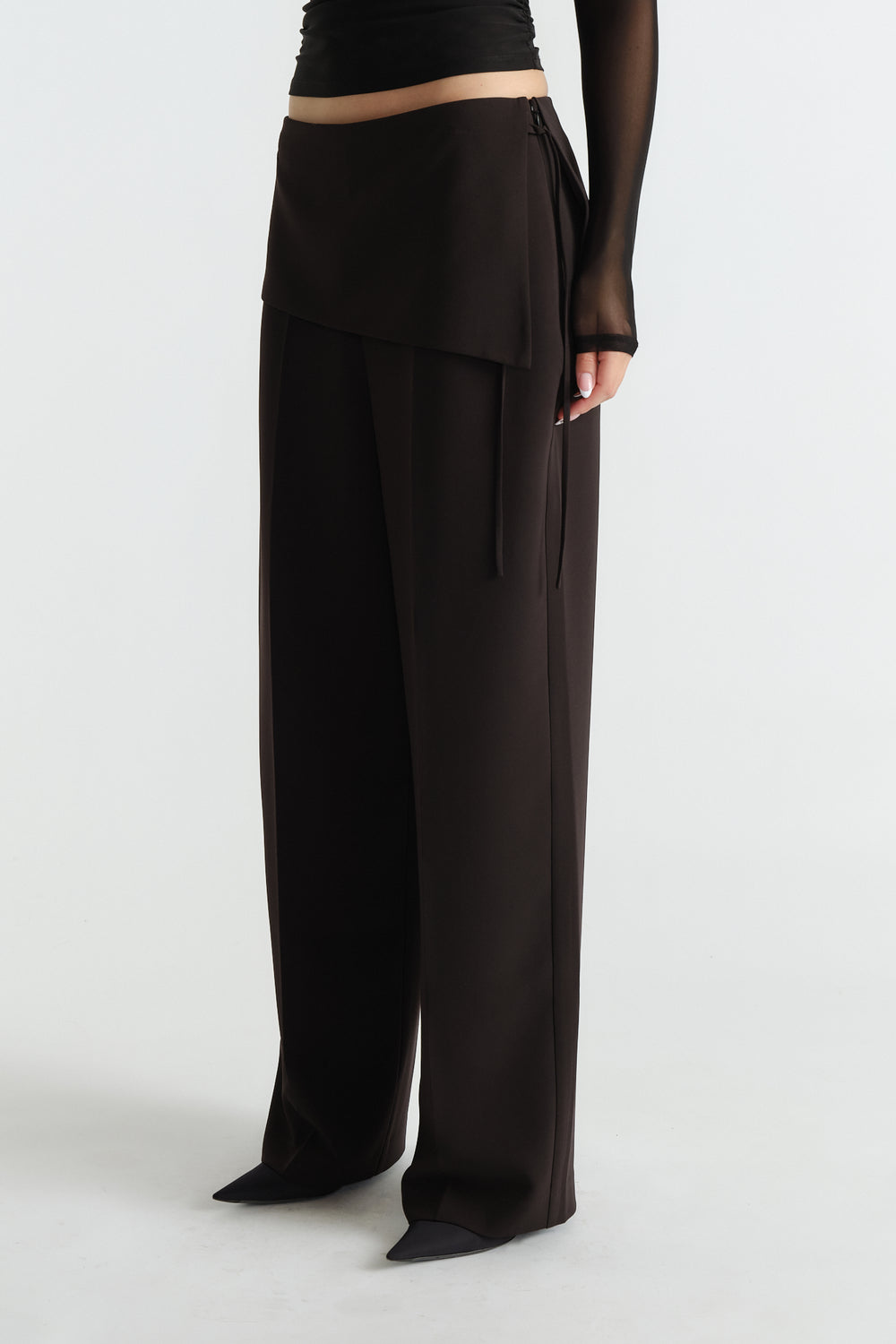 Side view of black tailored wide-leg pants with foldover detail worn with sheer long sleeve top and pointed shoes.