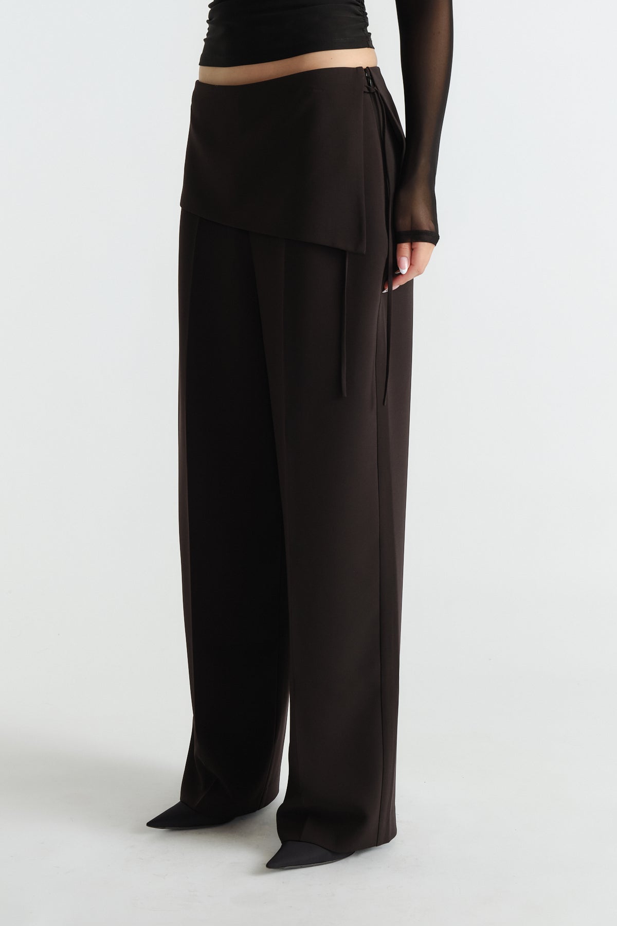 Side view of black tailored wide-leg pants with foldover detail worn with sheer long sleeve top and pointed shoes.