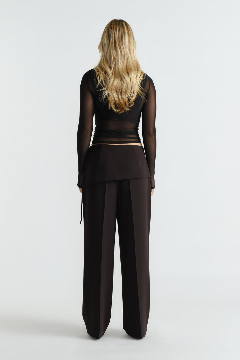 Back view of woman wearing tailored black foldover pants paired with a sheer long-sleeve black top and loose waves.