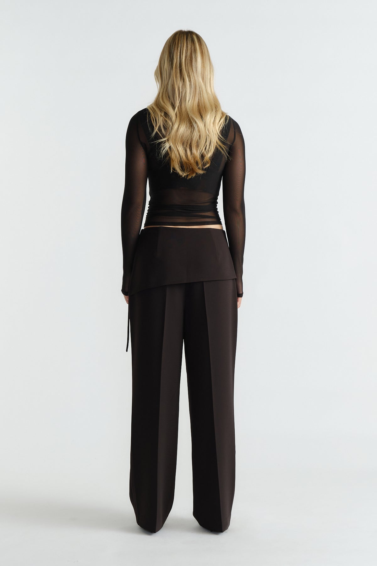 Back view of woman wearing tailored black foldover pants paired with a sheer long-sleeve black top and loose waves.