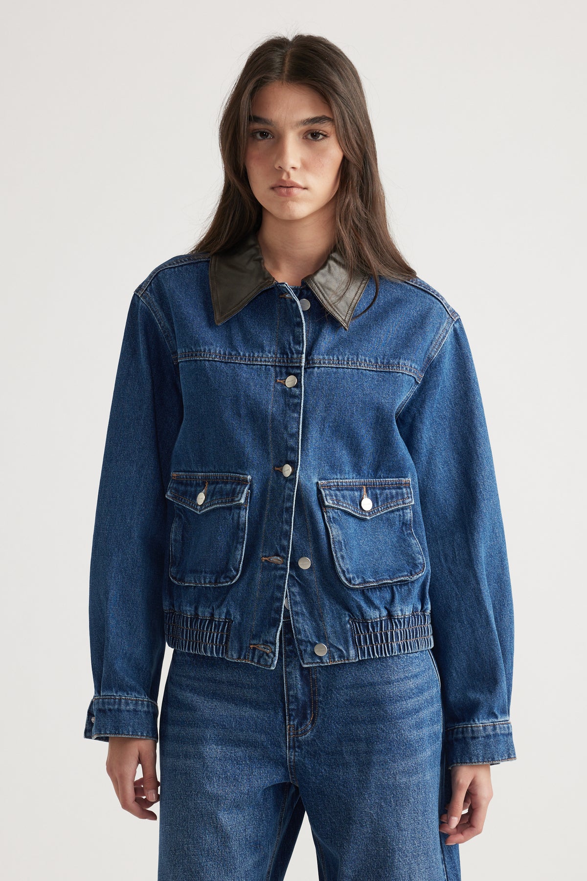 Woman wearing a blue denim jacket with contrast collar and front flap pockets paired with matching denim jeans against plain background