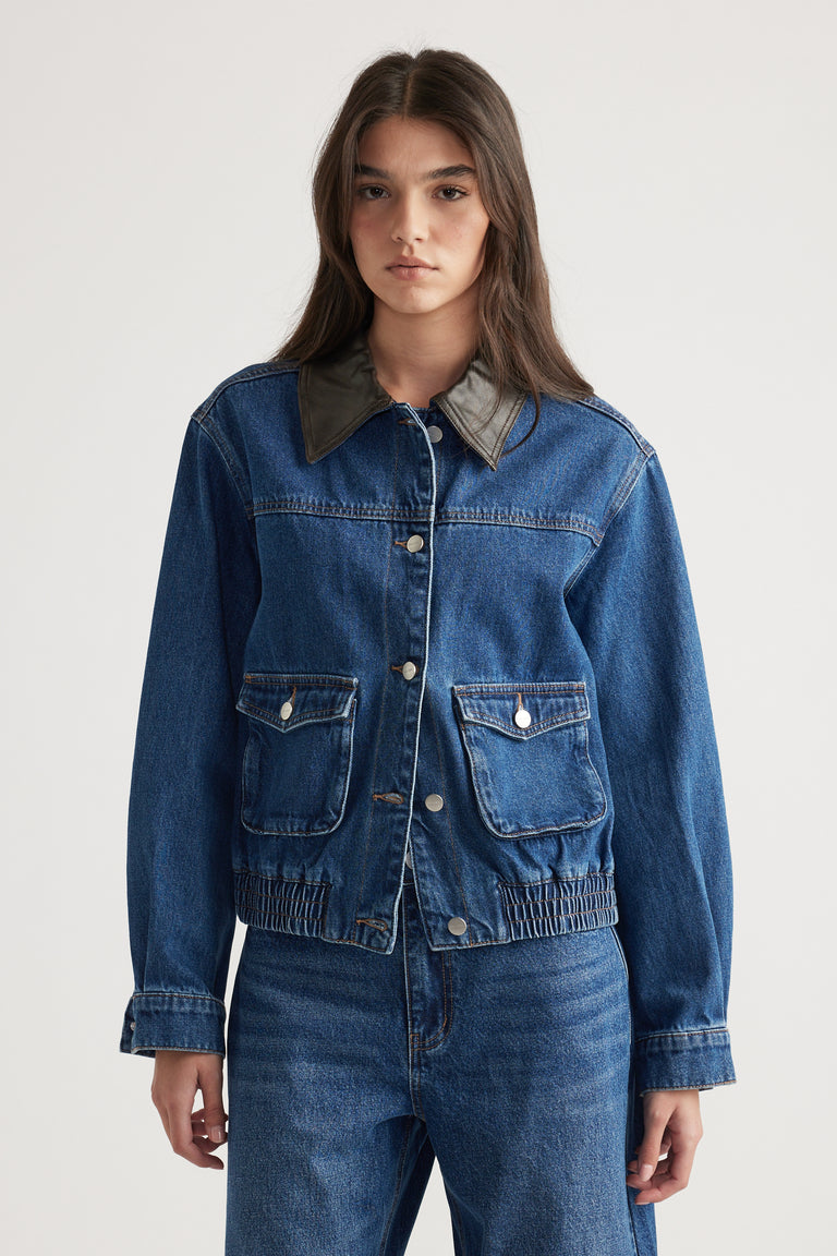 Woman wearing a blue denim jacket with contrast collar and front flap pockets paired with matching denim jeans against plain background