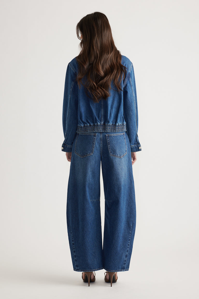 Woman seen from behind wearing a relaxed-fit denim jacket and wide-leg jeans against a plain backdrop.