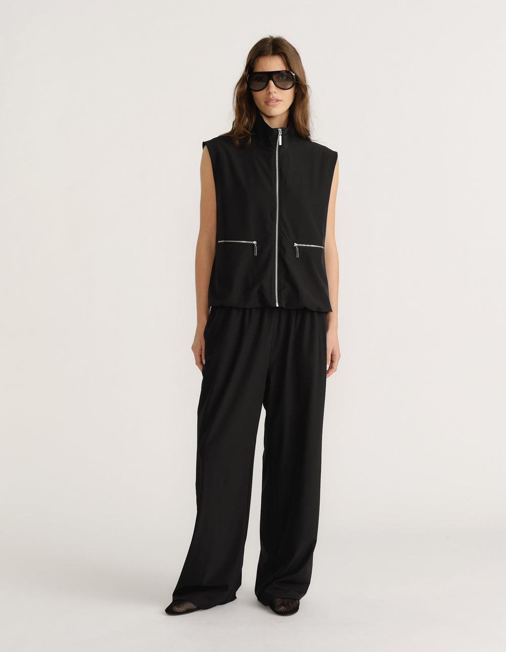 Woman wearing sleeveless black zip-up gilet with wide-leg matching black pants and oversized sunglasses