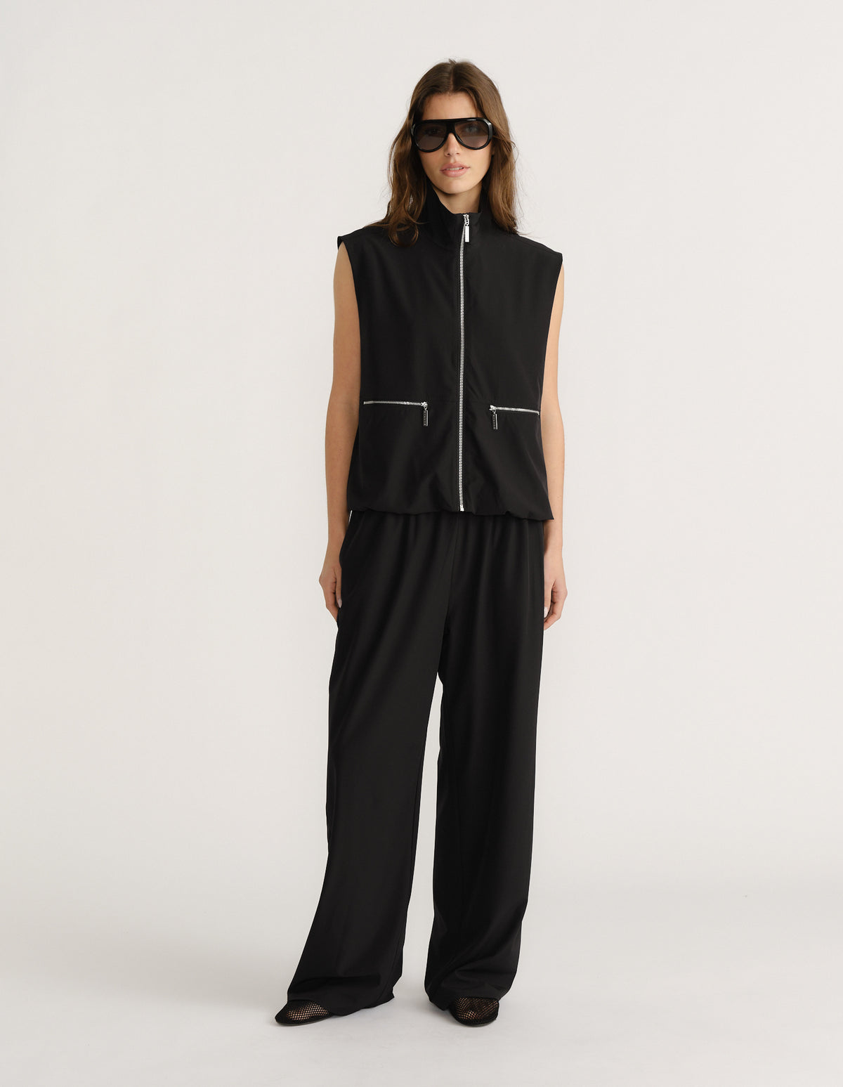 Woman wearing sleeveless black zip-up gilet with wide-leg matching black pants and oversized sunglasses