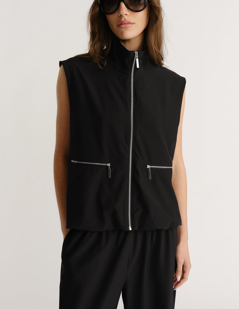 Woman wearing sleeveless black zip-up vest with high neck and zippered front pockets paired with loose black pants