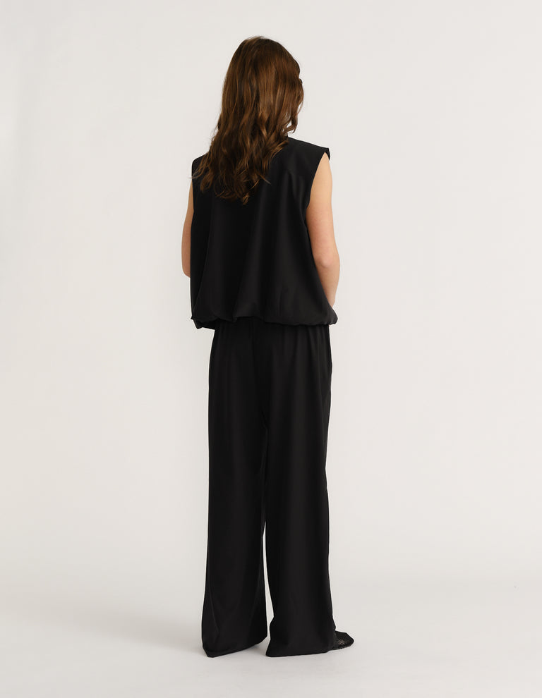 Woman wearing a sleeveless black top with wide-leg black pants, viewed from behind against a plain background