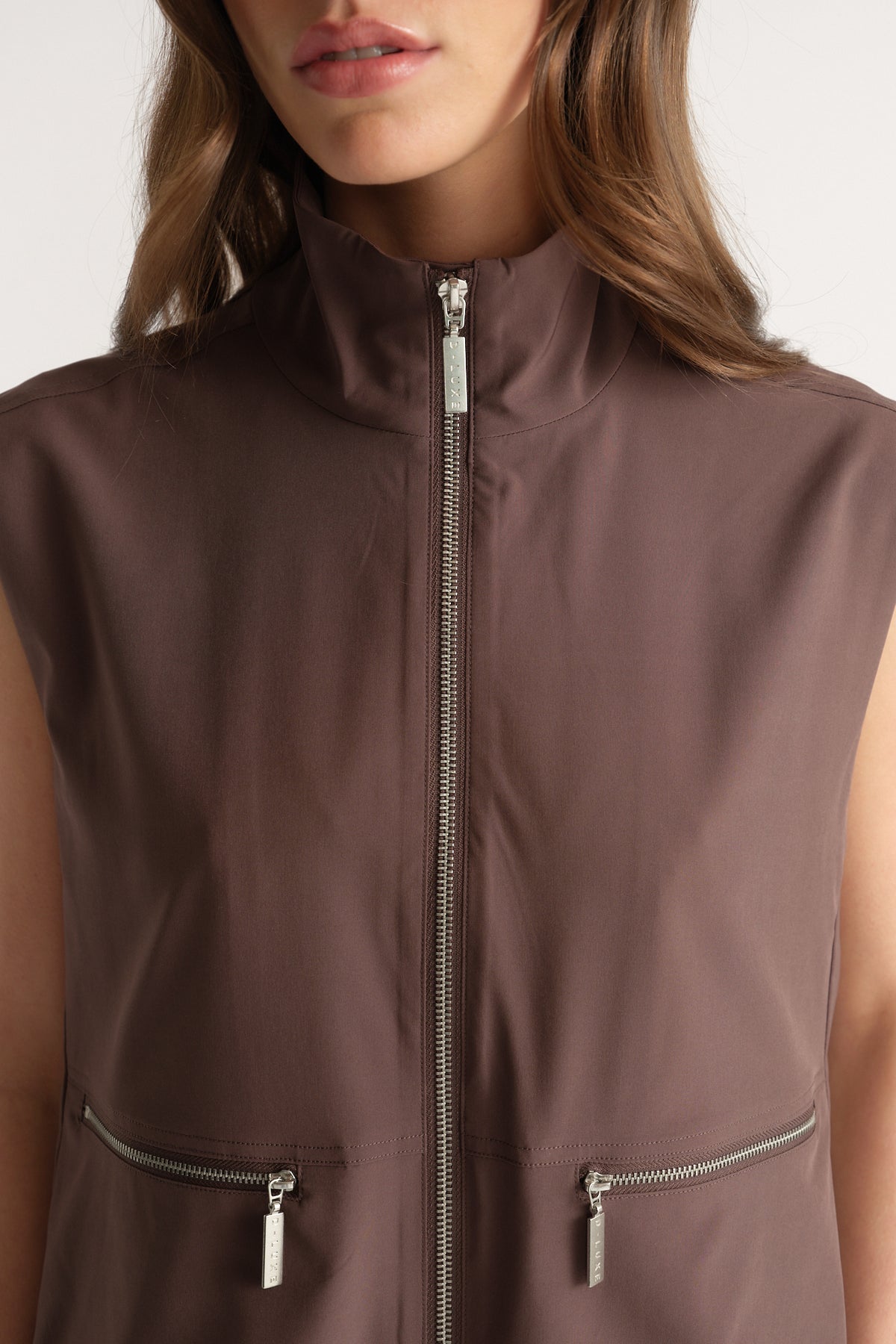 Woman wearing a brown sleeveless high neck gilet with a front zip closure and two zip pockets