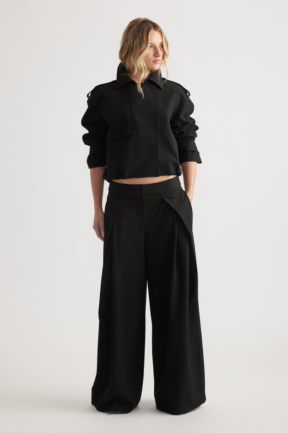 Woman wearing black wide leg pleated pants paired with a cropped jacket, hands in pockets, standing against white background