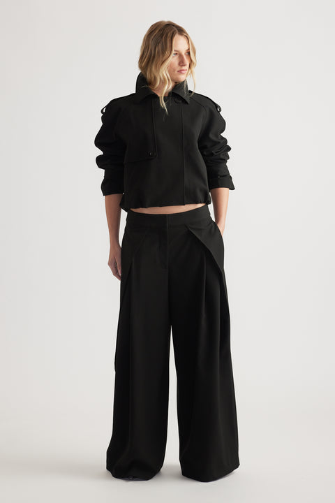 Woman wearing black wide leg pleated pants paired with a cropped jacket, hands in pockets, standing against white background