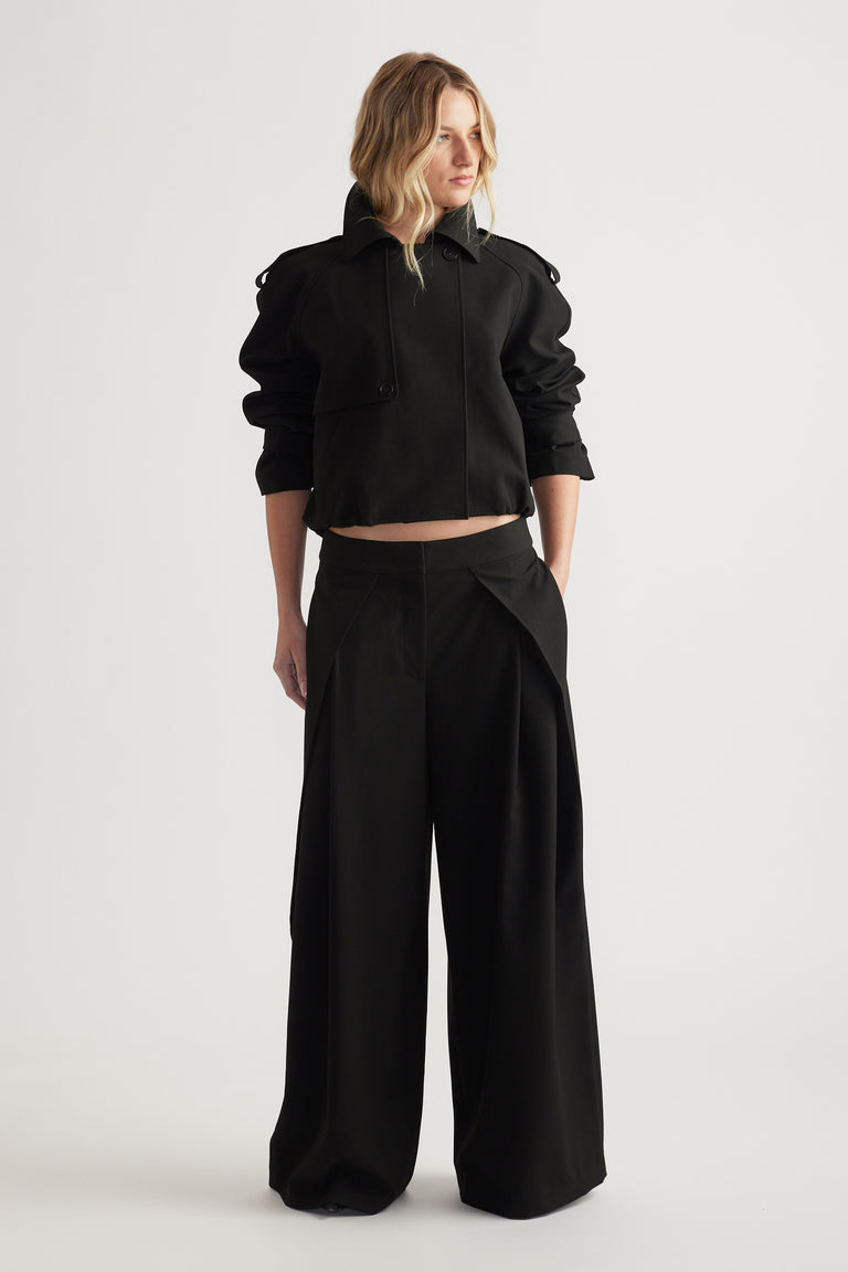 Woman wearing black wide leg pleated pants paired with a cropped jacket, hands in pockets, standing against white background