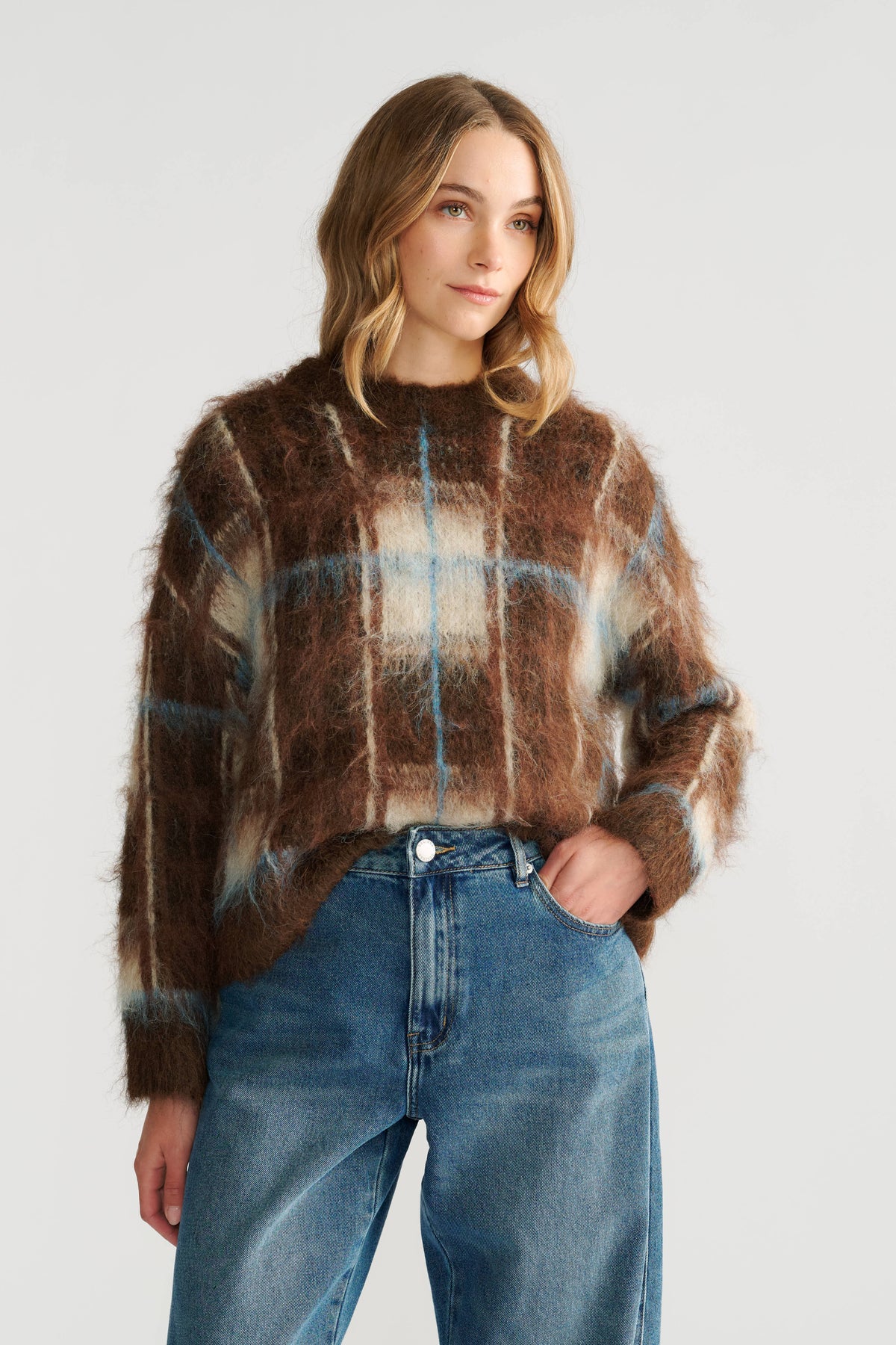 Woman wearing a cozy brown plaid knit jumper paired with relaxed blue high-waisted jeans against a neutral backdrop