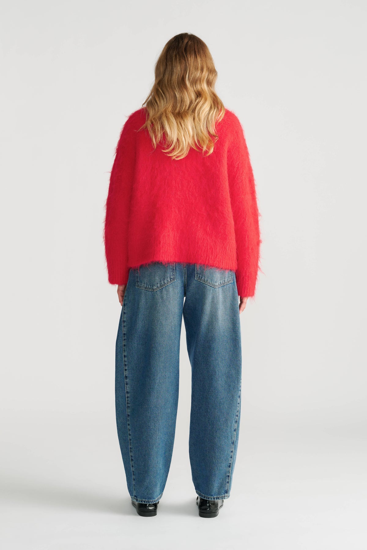 Woman wearing a vibrant red fuzzy knit jumper paired with relaxed fit blue jeans and black shoes, back view.