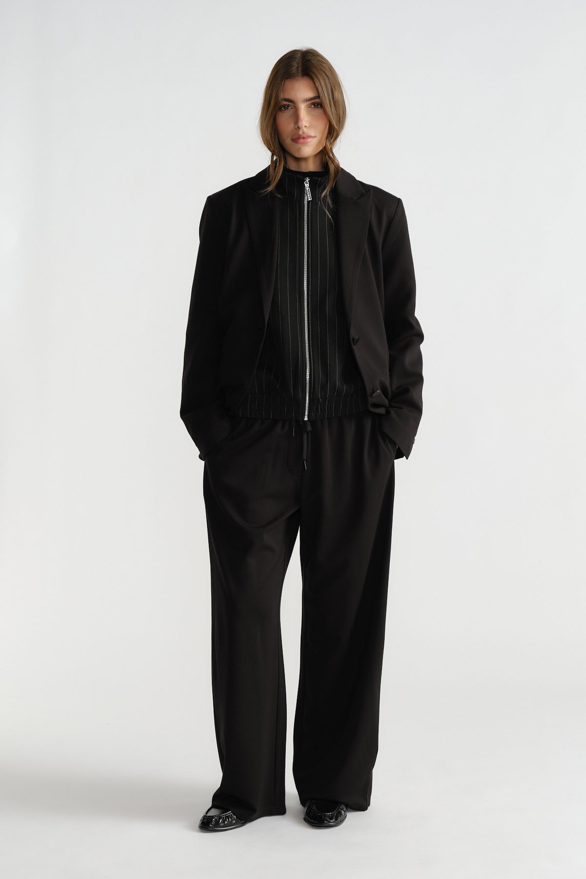 Woman wearing a relaxed-fit black blazer over a zip-front top, paired with wide-leg black trousers and black shoes