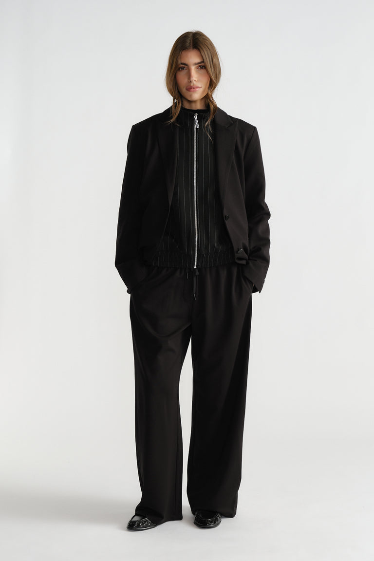 Woman wearing a relaxed-fit black blazer over a zip-front top, paired with wide-leg black trousers and black shoes
