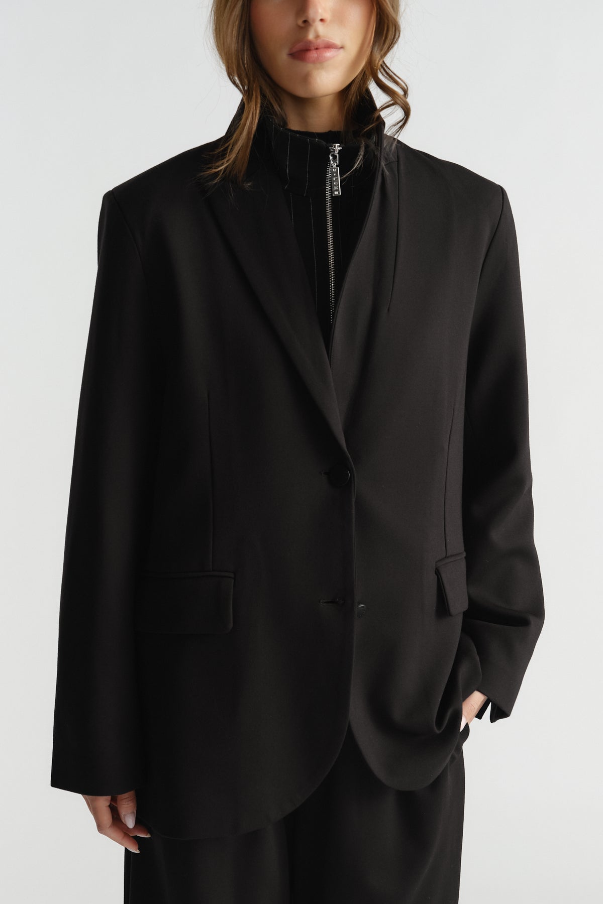 Woman wearing an oversized black blazer with notch lapels and flap pockets layered over a zip-up top