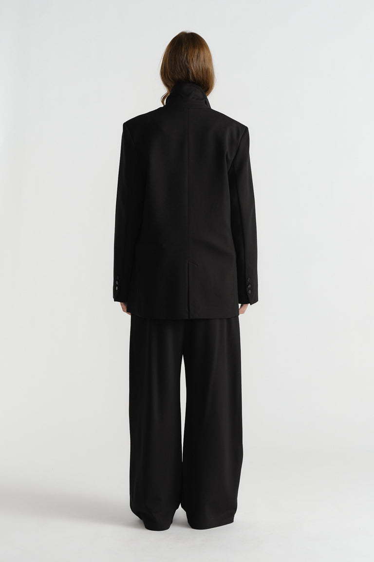 Back view of a woman wearing a structured black blazer with padded shoulders and matching wide-leg trousers