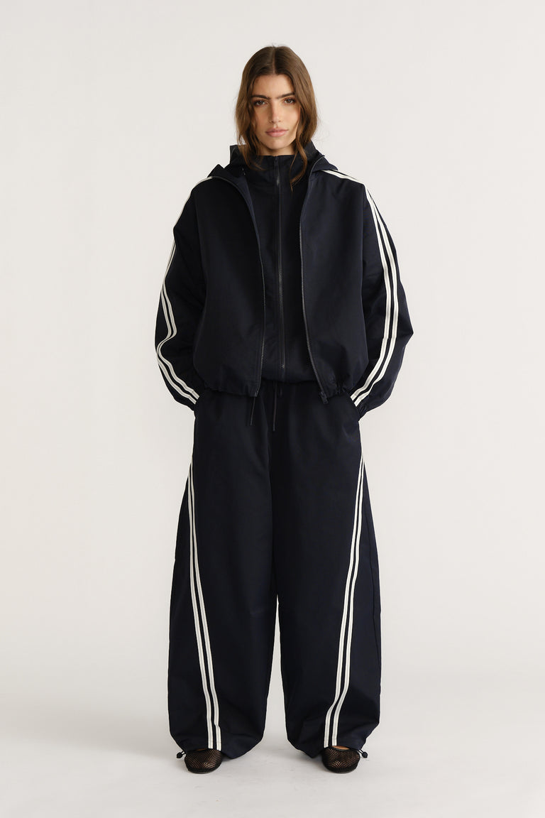 Model wearing navy relaxed fit pants with white side stripes paired with a matching sporty zip-up jacket for a chic casual look