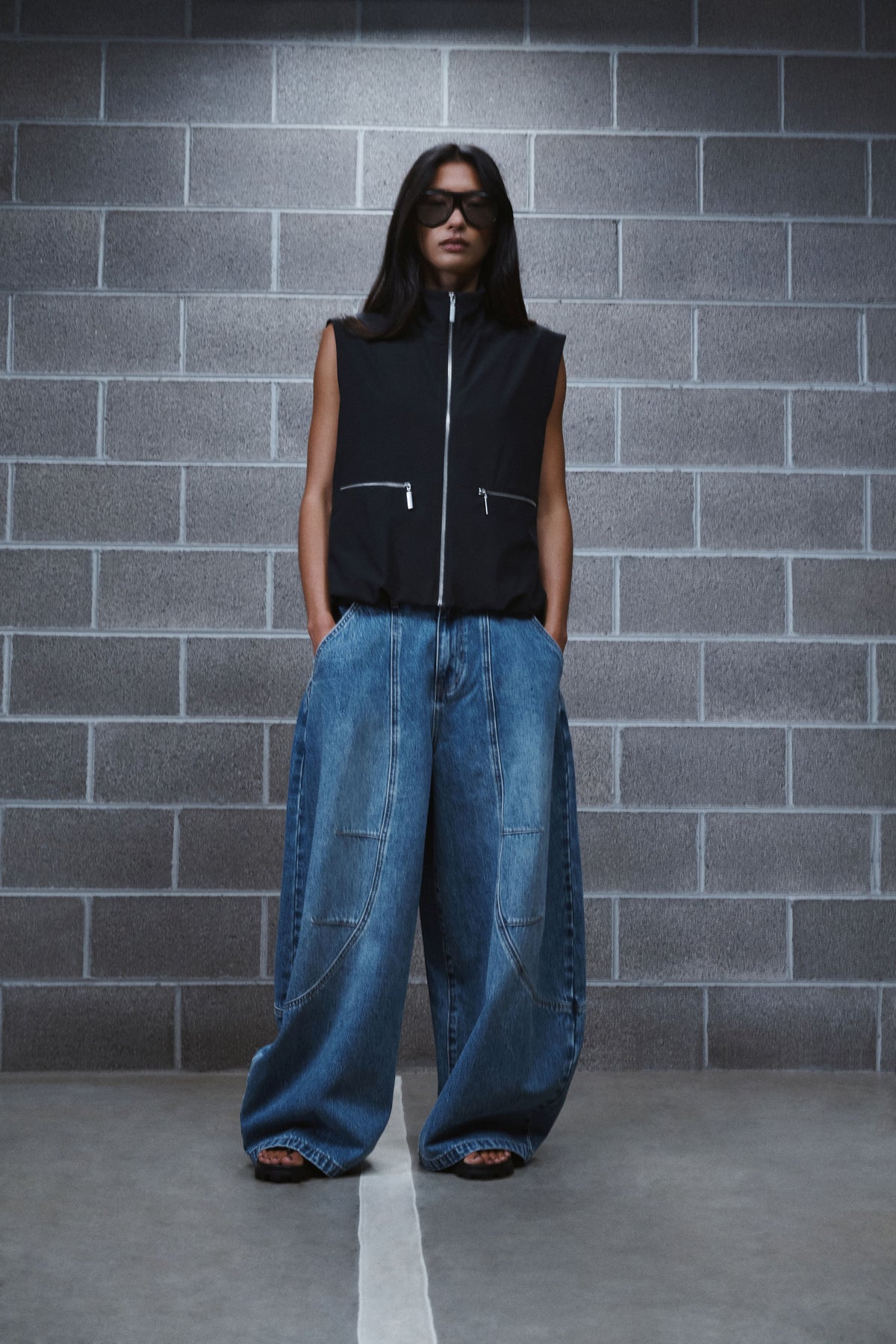 Woman wearing blue horseshoe jeans with a black sleeveless zip vest and sunglasses