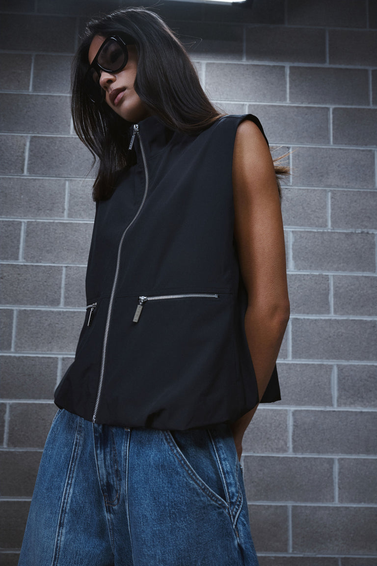 Woman wearing a black sleeveless high neck gilet with zip pockets, blue jeans and sunglasses