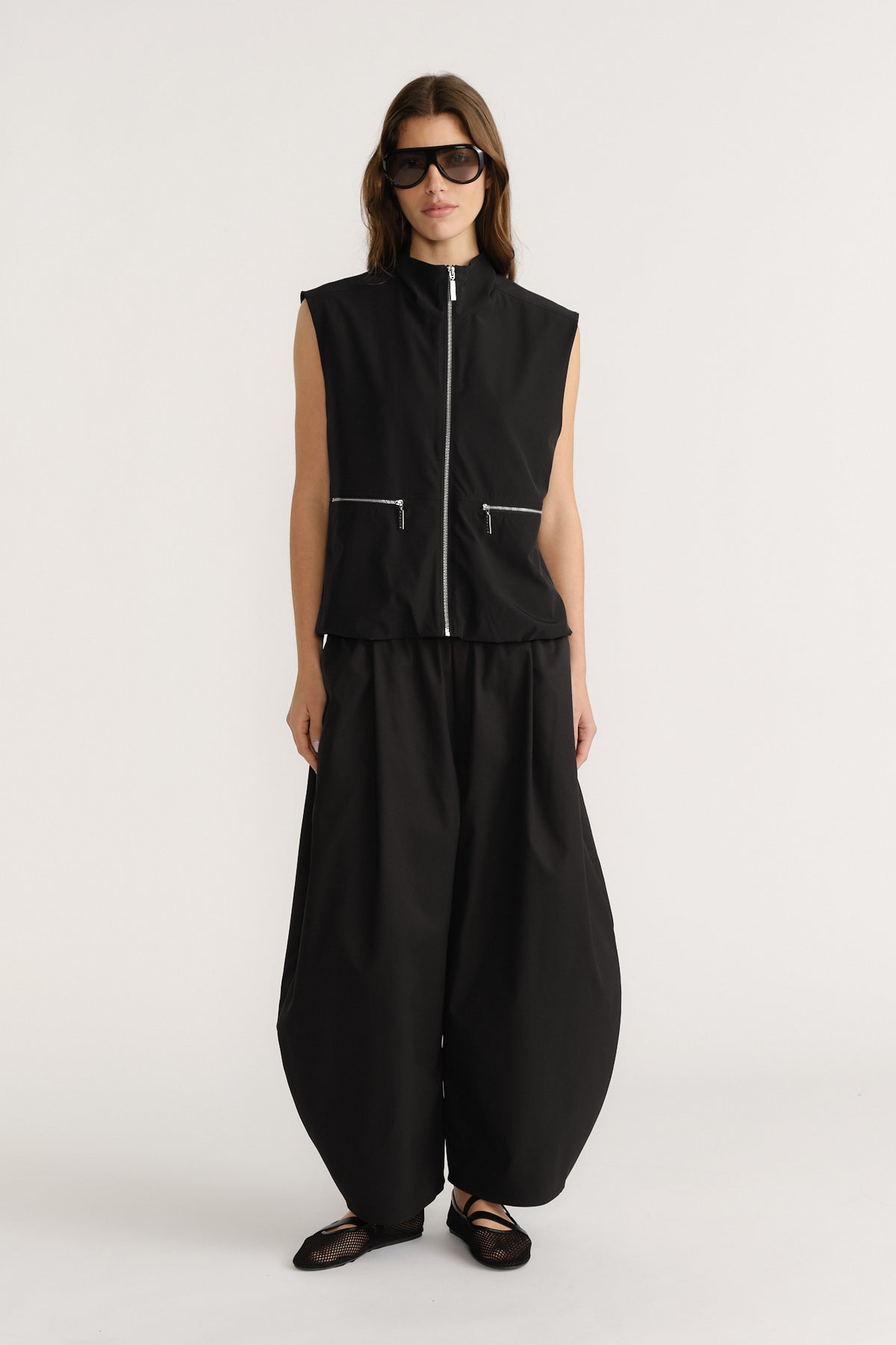 Woman wearing sleeveless black zip front top with zippered pockets paired with loose black barrel-leg pants and flat shoes