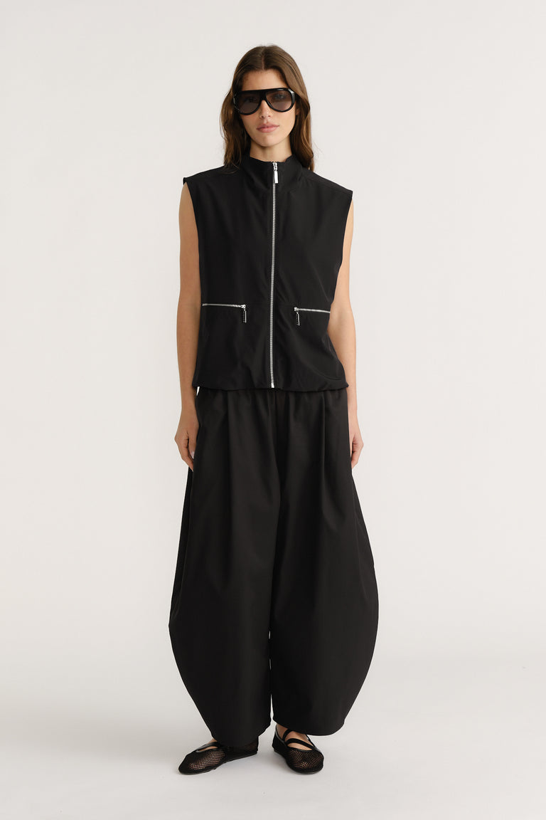 Woman wearing sleeveless black zip front top with zippered pockets paired with loose black barrel-leg pants and flat shoes