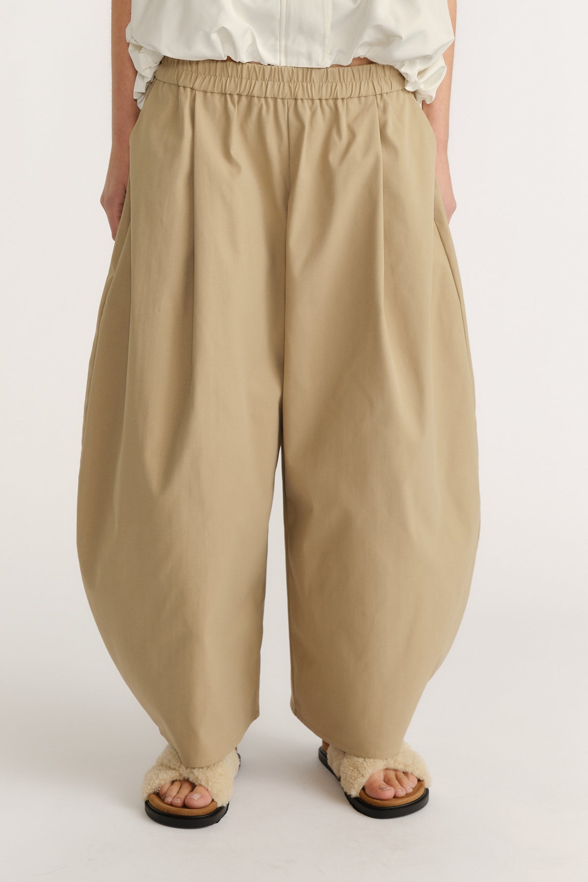 Loose-fit beige pants with a gathered waistband, side pockets, and a relaxed, modern silhouette paired with fuzzy sandals.