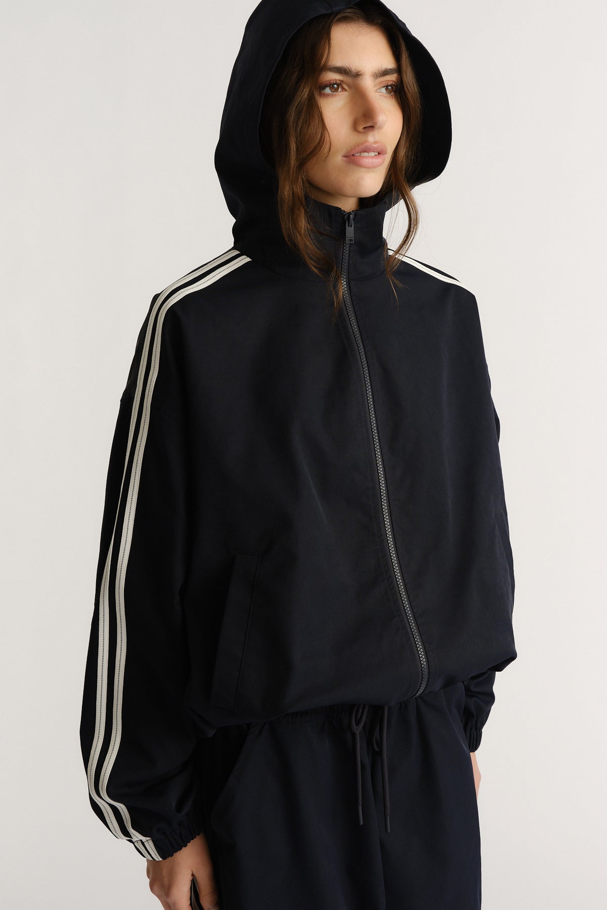 Woman wearing a navy hooded jacket with white side stripes and elasticated cuffs, styled for a laid-back sporty look