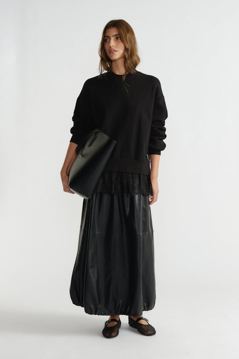Woman wearing a black lace-trimmed sweatshirt paired with a long leather skirt and carrying a large black tote bag.