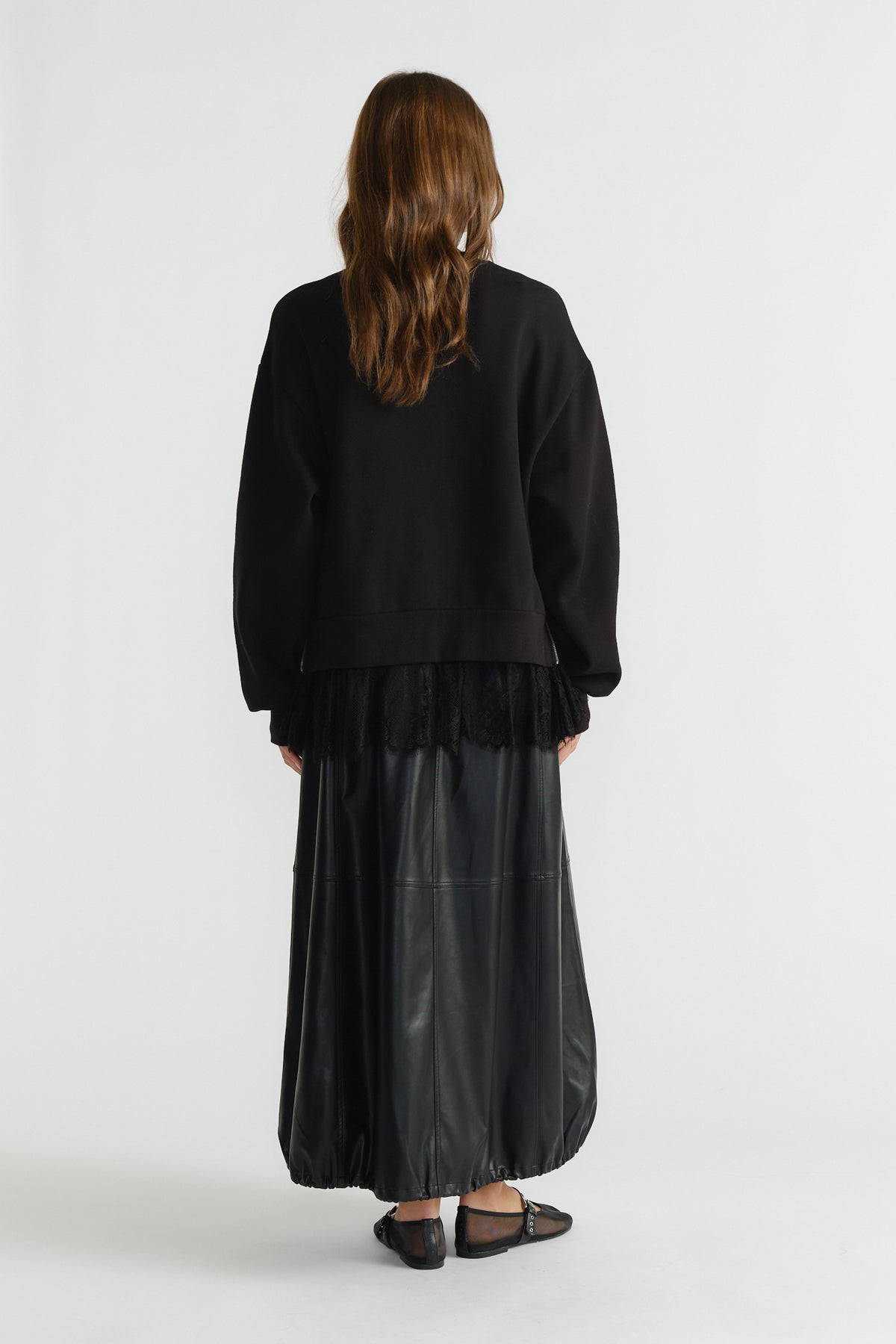 Back view of woman wearing black sweatshirt layered over lace hem with a long black faux leather skirt and black flats.