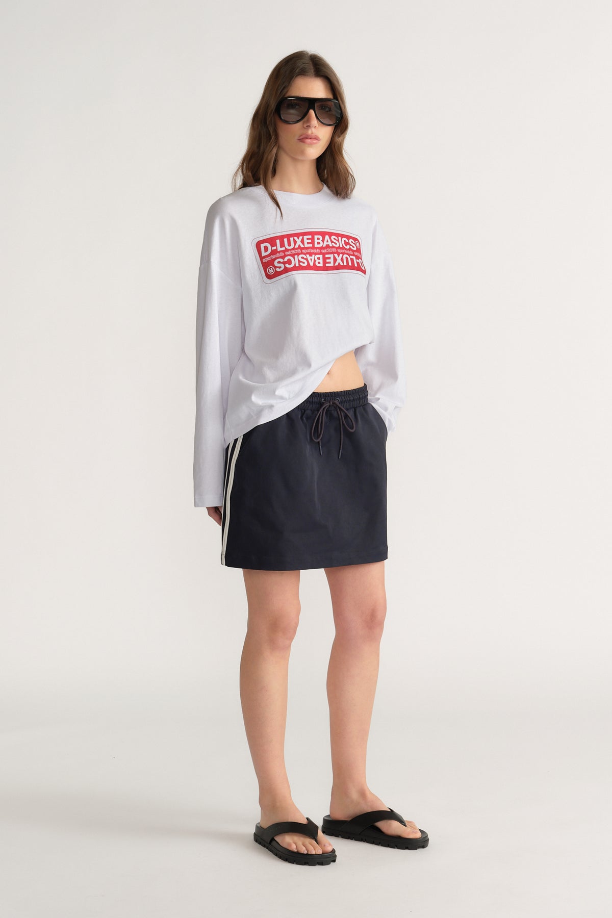 Woman wearing a white graphic long sleeve tee with a black side stripe mini skirt, sunglasses and sandals