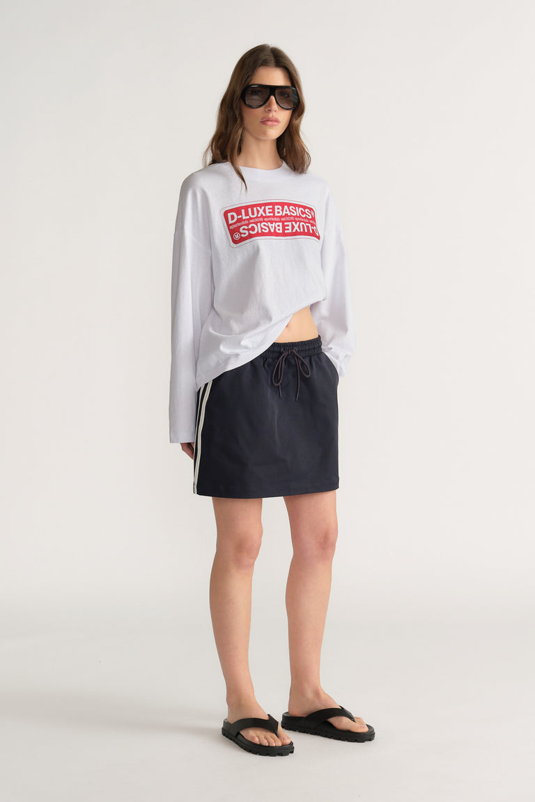 Woman wearing a white graphic long sleeve tee with a black side stripe mini skirt, sunglasses and sandals