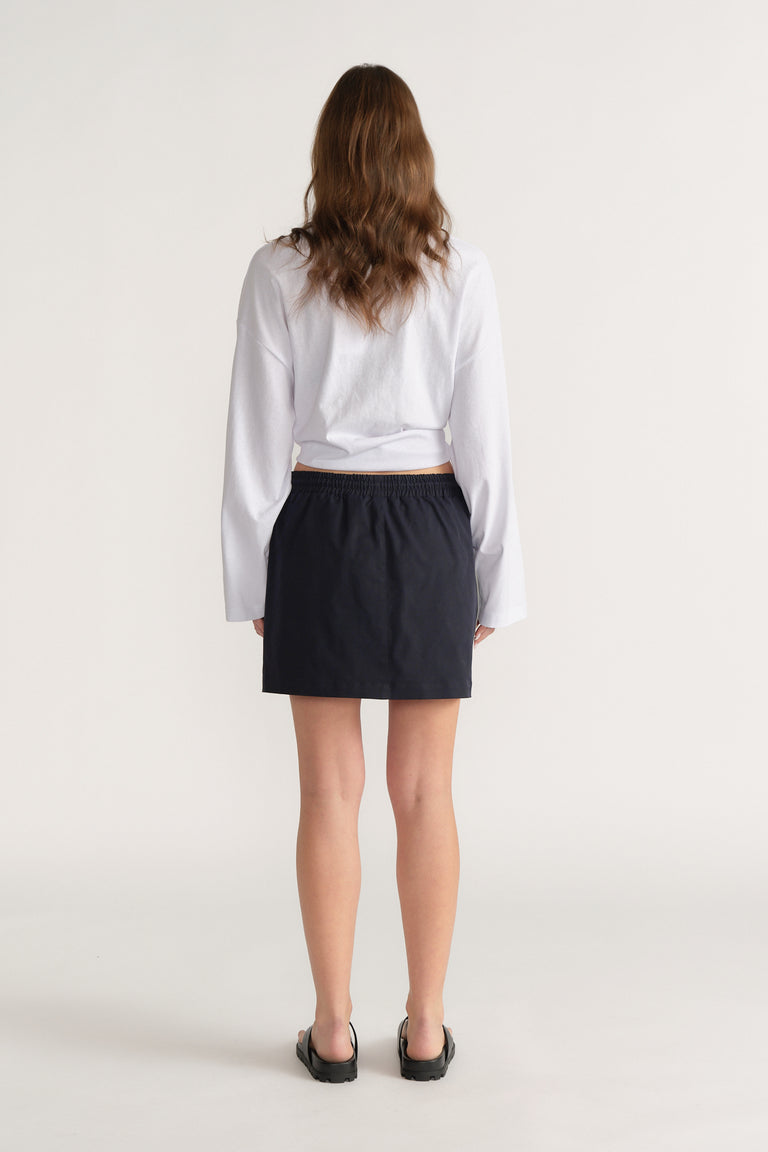 Model viewed from behind wearing a black elastic waist mini skirt, white long sleeve shirt and black loafers