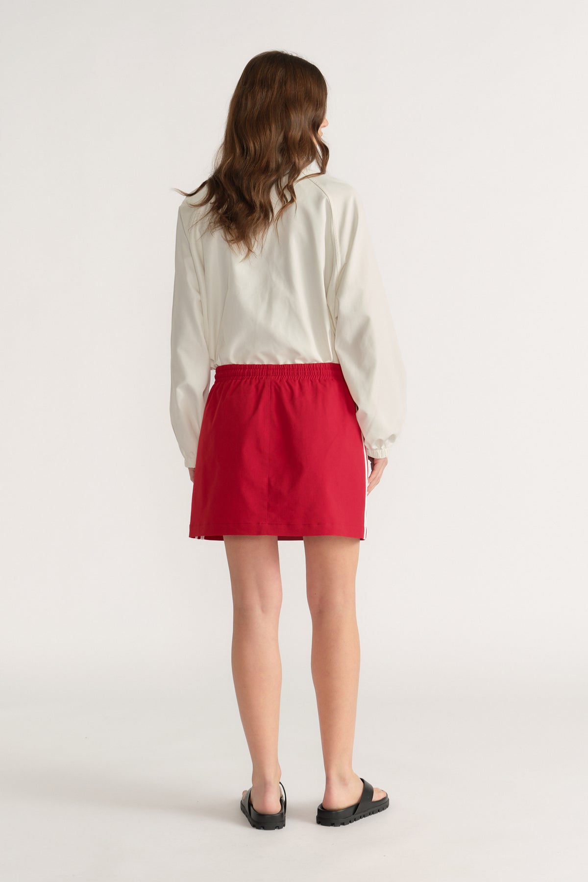 Woman viewed from behind wearing a red elastic waist mini skirt, cream long sleeve blouse and black sandals
