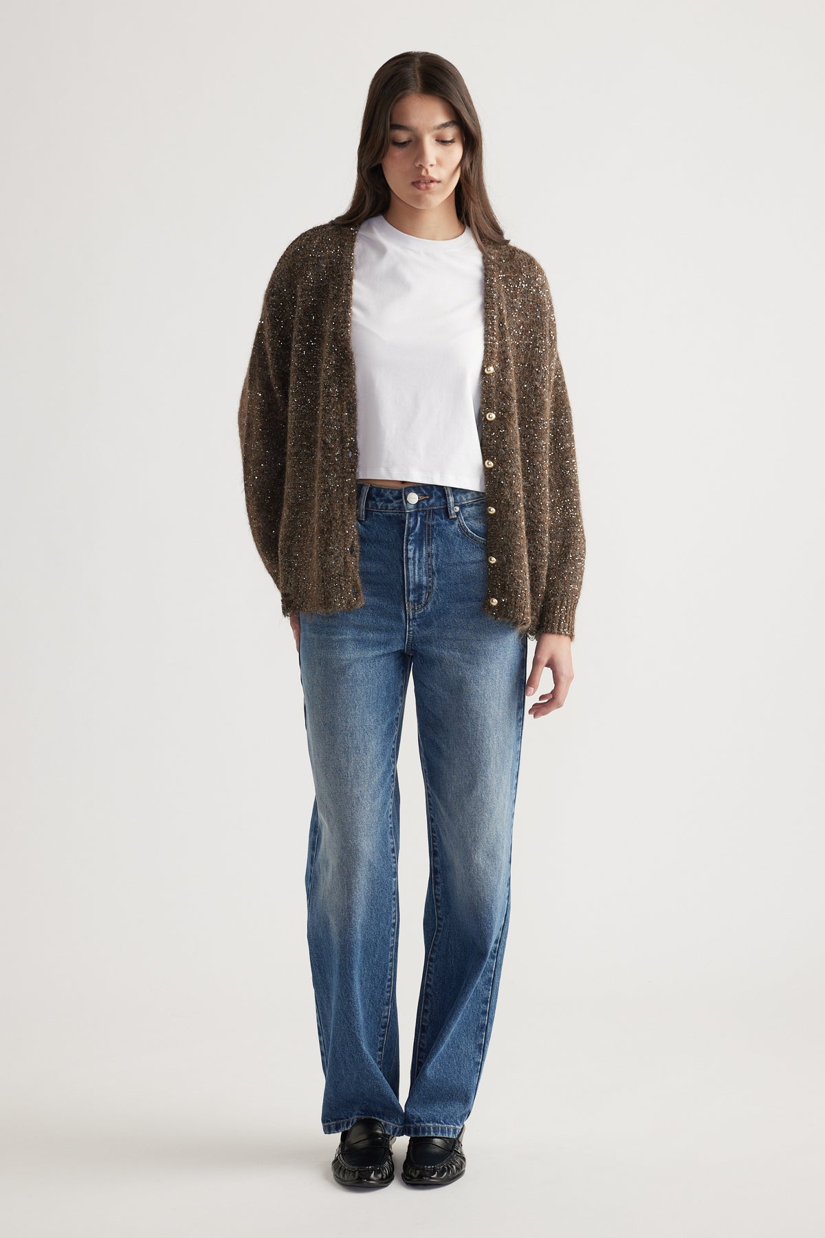 Woman wearing a bronze sequined cardigan over a white tee with relaxed blue denim jeans and black shoes
