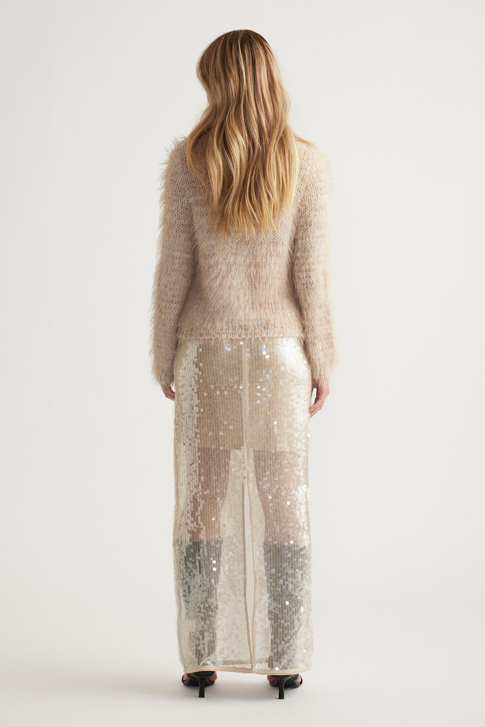 Back view of woman wearing a soft beige fluffy knit jumper paired with a long sheer sequin skirt and heels