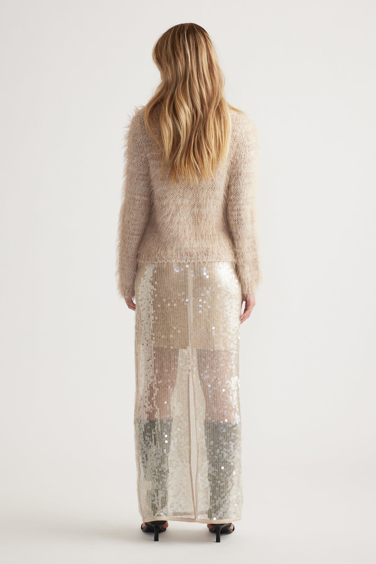 Back view of woman wearing a soft beige fluffy knit jumper paired with a long sheer sequin skirt and heels