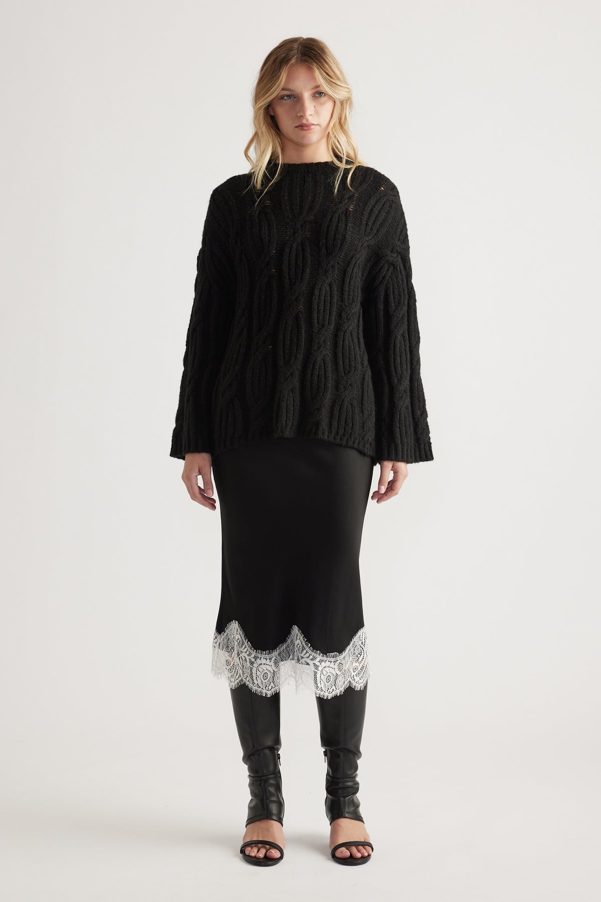 Woman wearing a loose black cable knit jumper paired with a black skirt featuring white lace trim and black boots.
