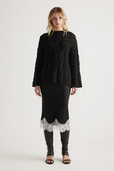 Woman wearing a loose black cable knit jumper paired with a black skirt featuring white lace trim and black boots.