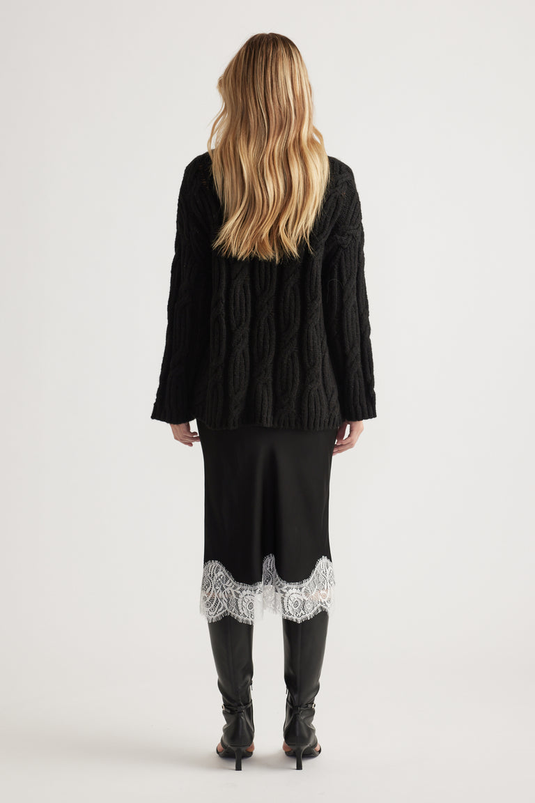 Woman wearing a black textured cable knit jumper paired with a black midi skirt featuring white lace trim and black boots