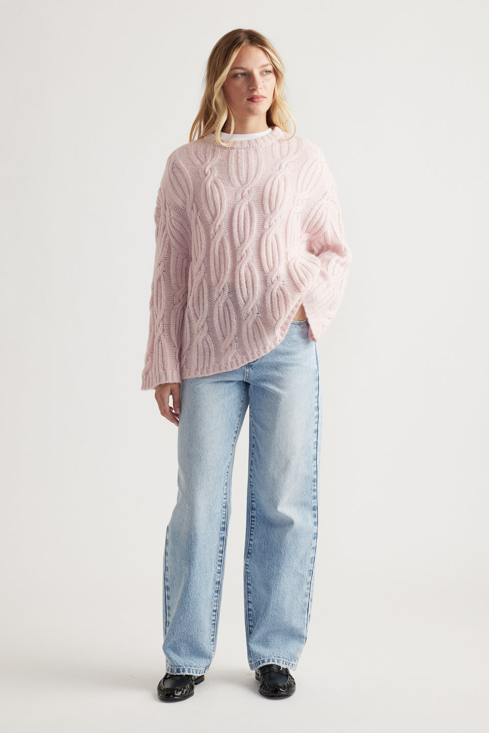 Woman wearing a soft pink cable knit jumper with relaxed light-wash jeans and black shoes in a clean studio setting