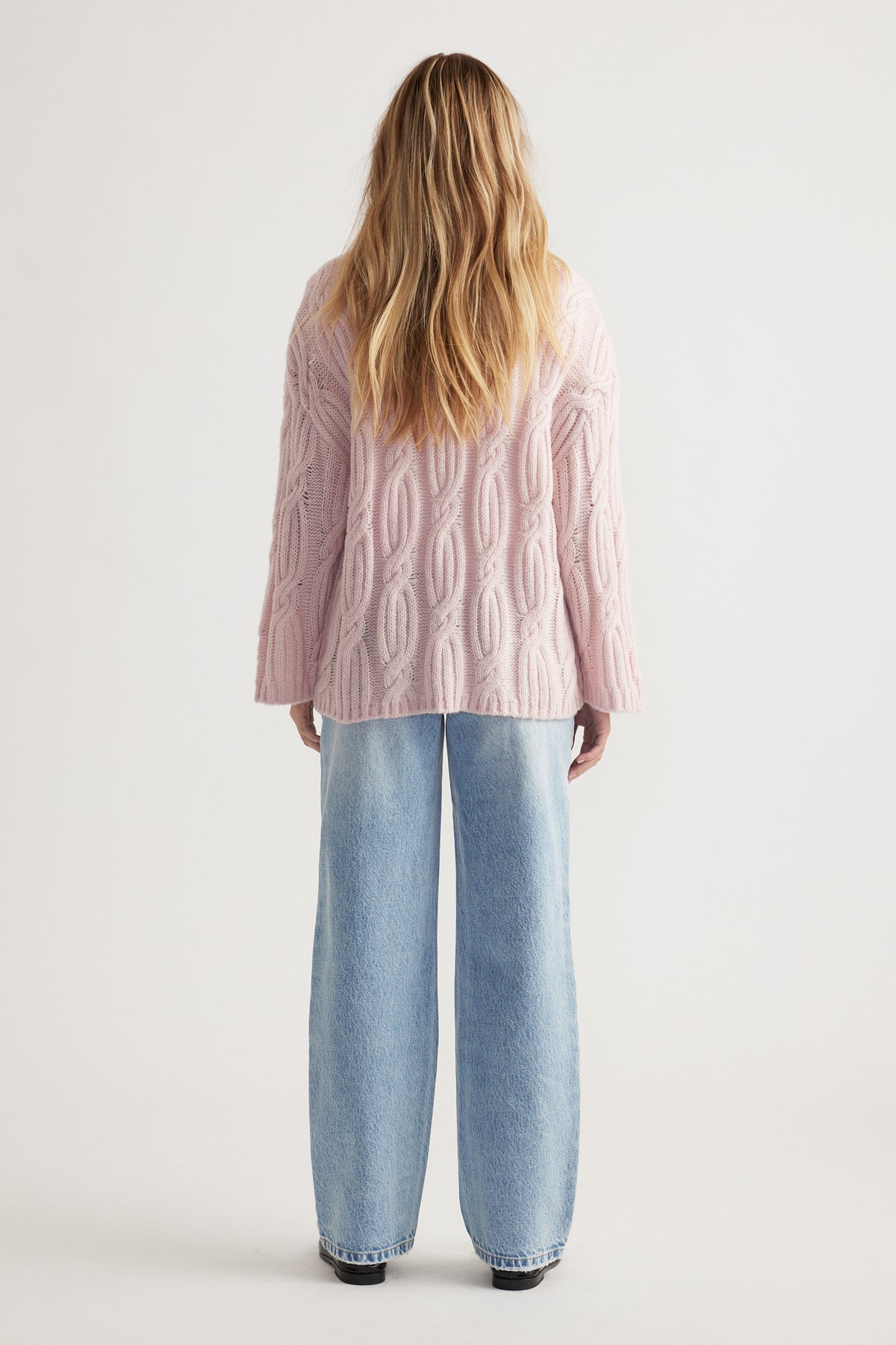 Woman with loose blonde hair wearing a soft pink cable knit jumper paired with light wash wide-leg jeans from behind