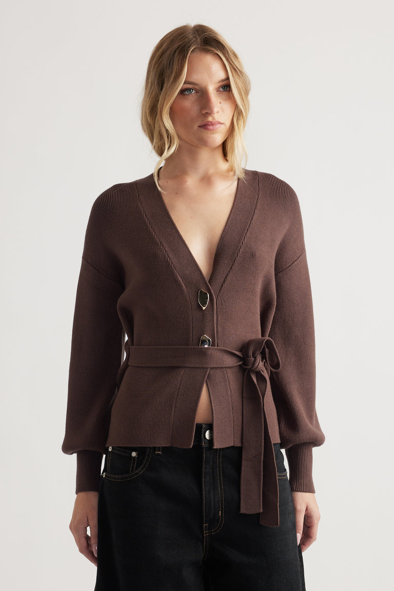 Woman wearing a brown knit cardigan with tie waist and long sleeves styled with high-waisted black jeans against a neutral backdrop