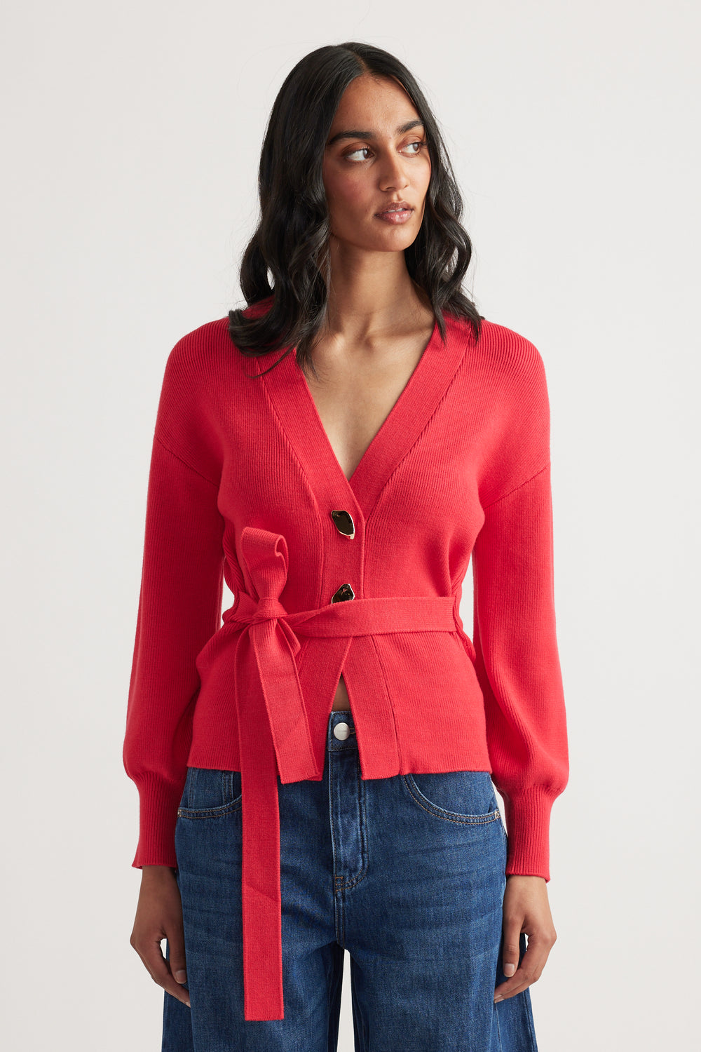 Woman wearing a bright red knit cardigan with a tie waist and large buttons, paired with blue denim jeans