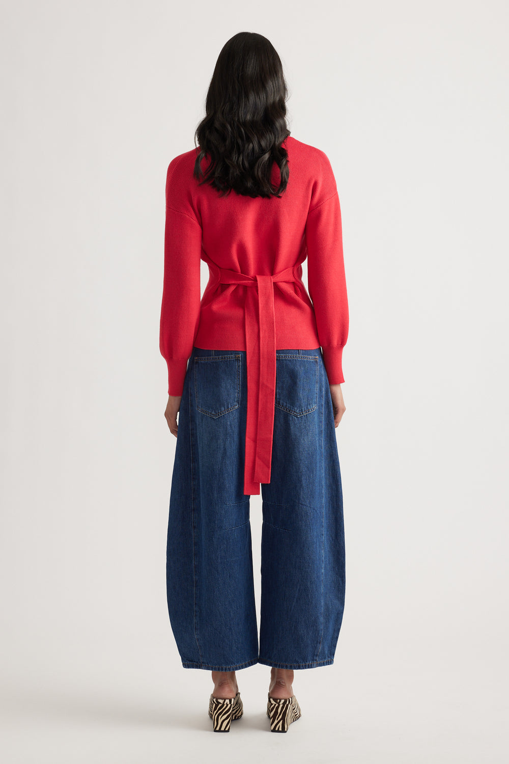 Woman wearing a bright red knit cardigan with a tie back detail paired with wide-leg blue denim jeans and animal print heels