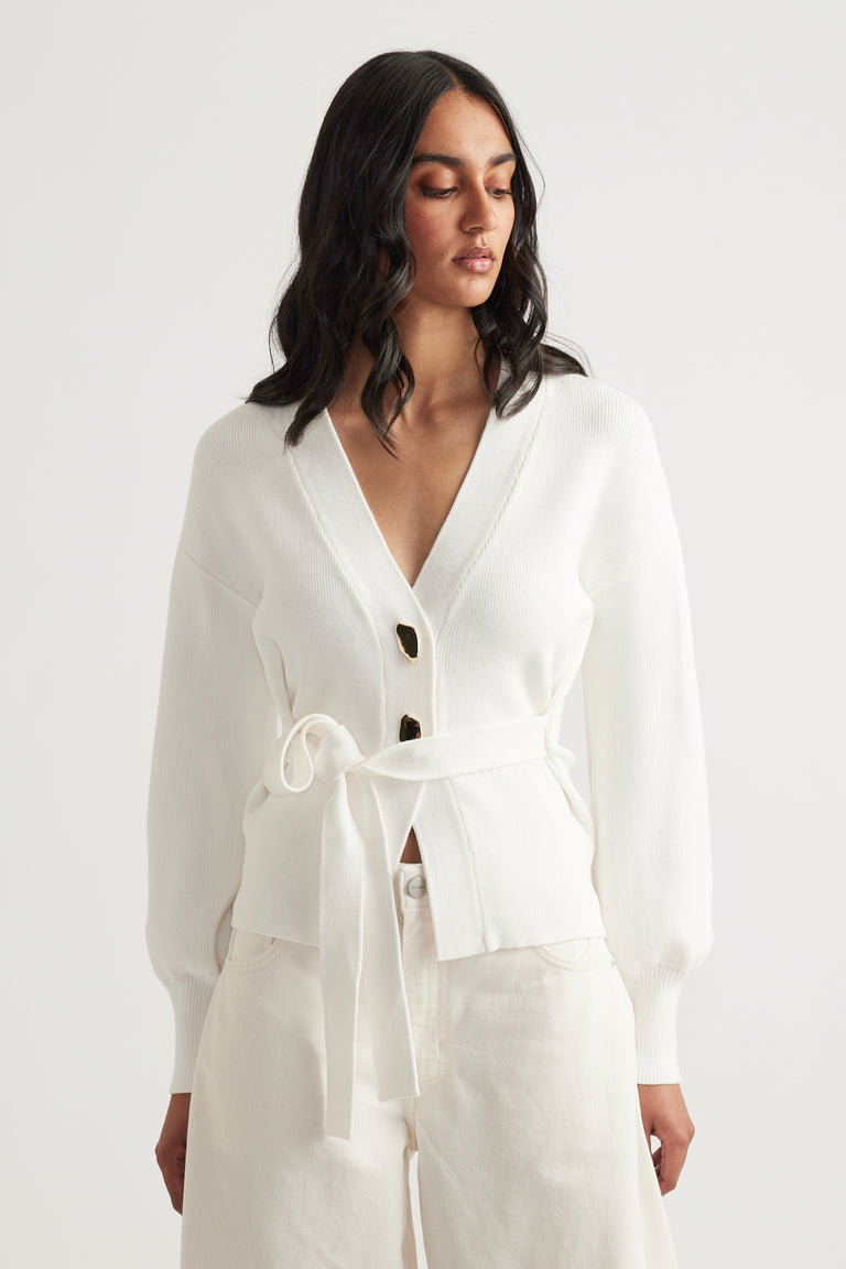 Woman wearing a white knit cardigan with a tie belt and dark buttons paired with loose ivory pants