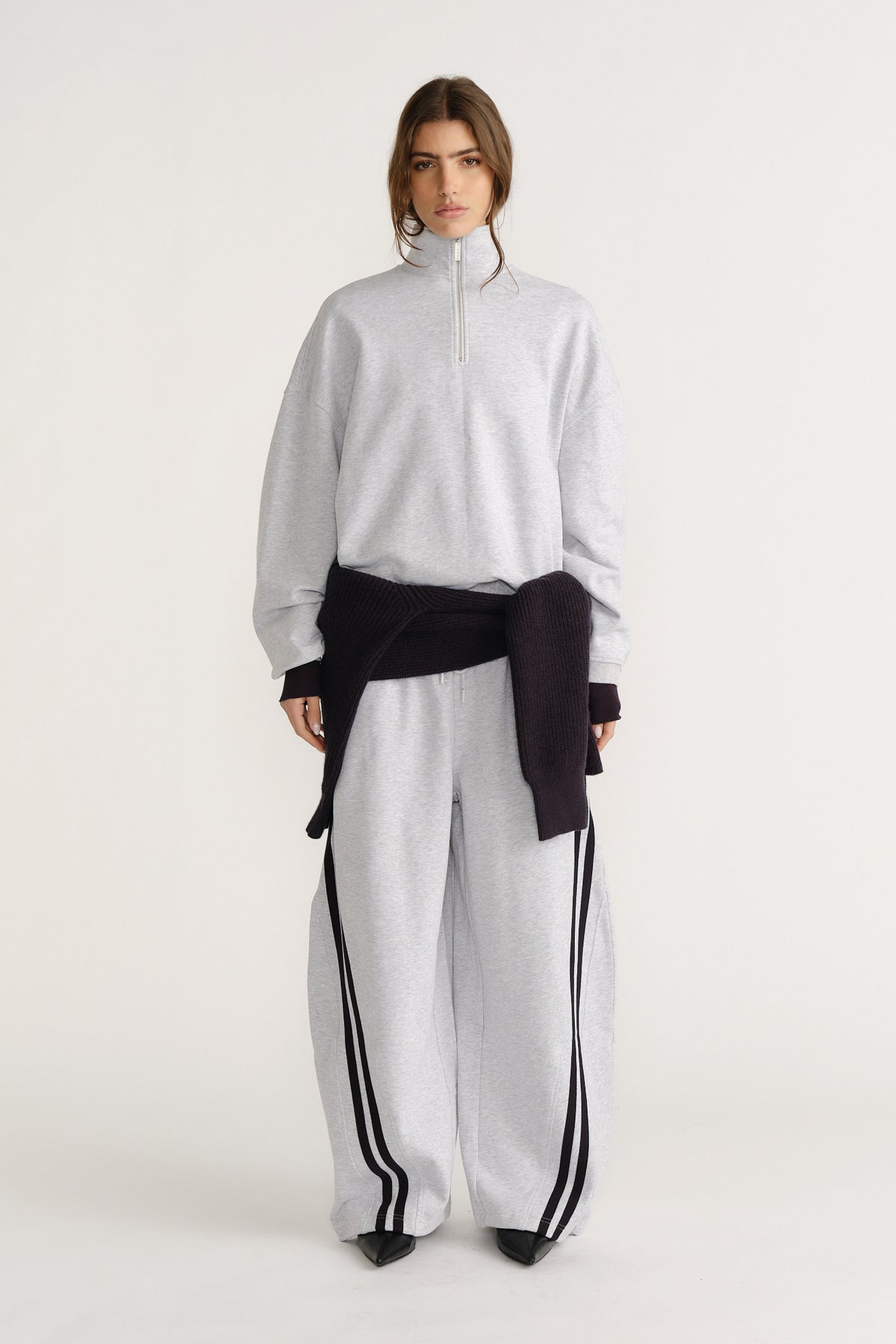 Woman wearing a light grey quarter-zip sweat paired with matching wide-leg pants featuring black side stripes and a black sweater tied at waist