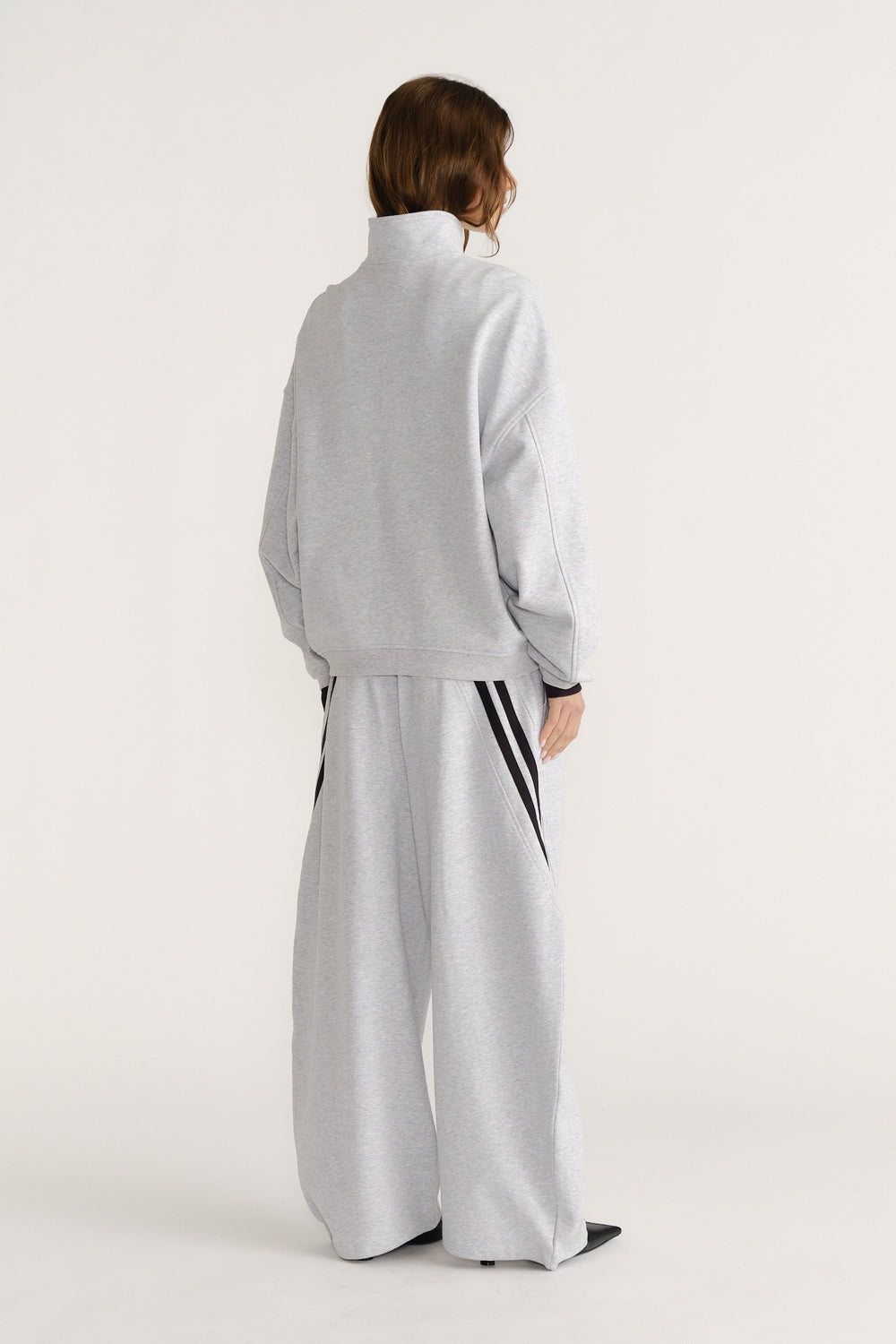 Woman seen from behind wearing a relaxed light grey quarter zip sweat with matching wide-leg pants featuring black side stripes